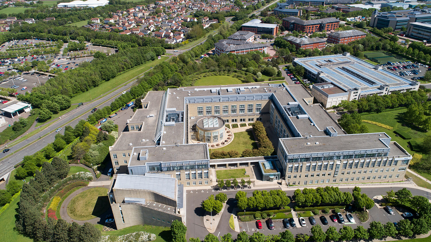 Drone photography of office campus at Edinburgh business park. You can see the modern building which is surrounded by trees and lawns. There is also a garden in the middle of the building. At the front is a parking area framed by trees. It is a bright sunny day. By Edinburgh Drone Photographers Blink Imaging
