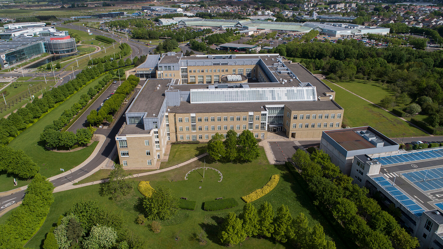 Aerial drone photography of large office building set in grounds with grass and trees. You can see solar panels on the roof of the building. In the distance you can see the Gyle shopping centre in the distance