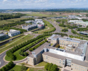 Drone photography of Edinburgh business park and the Gyle. You can see a large office building in the foreground surrounded by grass and trees. In the distance you can see the Gyle shopping centre, the runway of Edinburgh Airport and the Forth Bridges. By Edinburgh drone photographers Blink Imaging