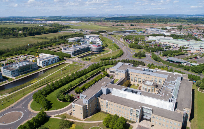 Drone photography of Edinburgh business park and the Gyle. You can see a large office building in the foreground surrounded by grass and trees. In the distance you can see the Gyle shopping centre, the runway of Edinburgh Airport and the Forth Bridges. By Edinburgh drone photographers Blink Imaging